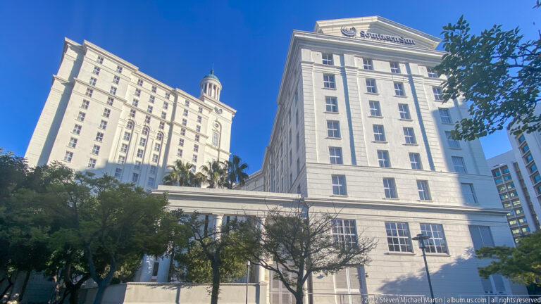 Two pale multi-story hotel towers connected by an enclosed elevated bridge, seen above roadside trees.
