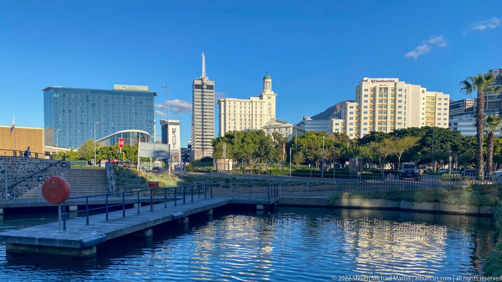 Calm canal basin with a low waterfront walkway and Cape Town hotel and convention-center skyline under a blue sky.