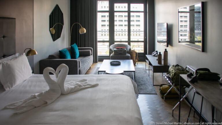 Wide hotel-room interior with bed and towel swans at left, sofa and coffee table center, and tall windows at the far end.