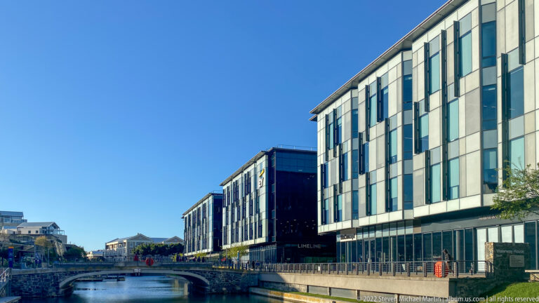 Modern multi-story buildings beside a narrow canal, with an arched bridge in the distance.