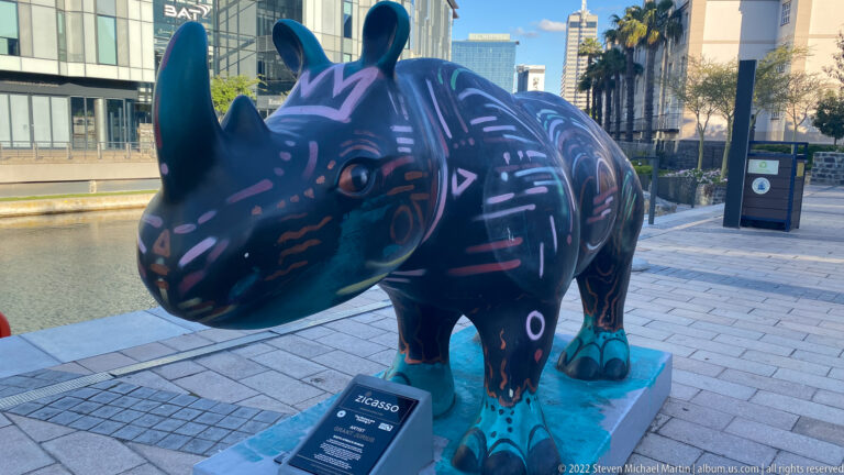 Dark painted rhinoceros sculpture with colorful graphic markings on a turquoise base beside the waterfront canal.
