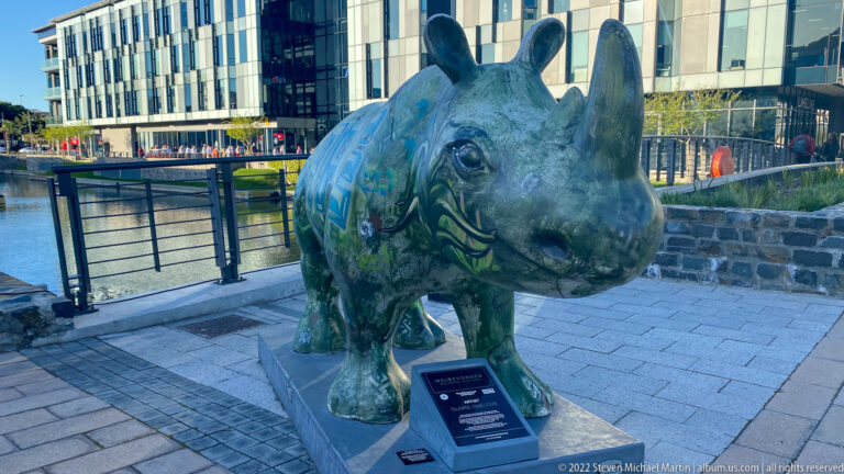 Green and blue painted rhinoceros sculpture beside the canal with a small interpretive plaque at its feet.