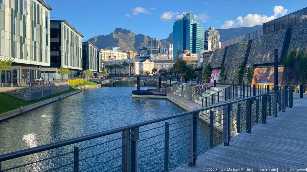 Boardwalk and railing beside a waterfront canal, with modern Cape Town buildings and Table Mountain in the distance.