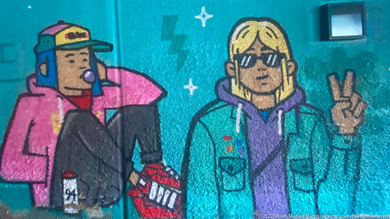 Turquoise mural with two cartoon figures, including a blond person in sunglasses making a peace sign.