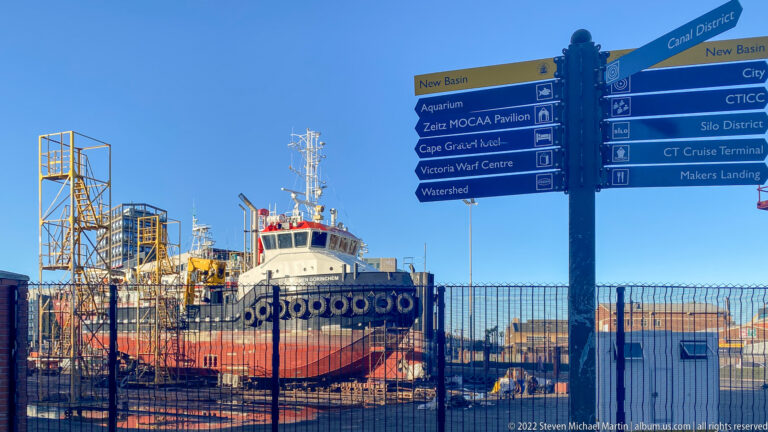 Red-and-white harbor workboat behind a fence and a multi-direction V&A Waterfront signpost.