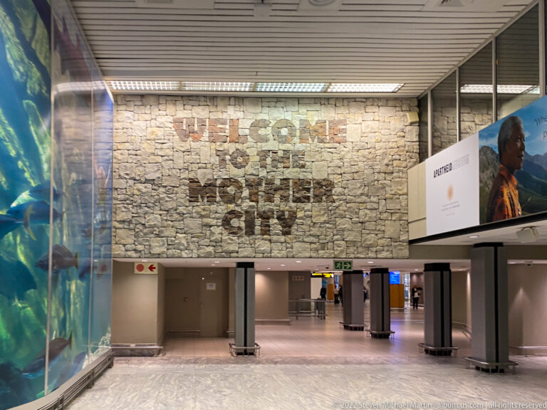 Airport concourse beneath a stone wall reading “WELCOME TO CAPE TOWN,” with large advertising panels and corridors below.