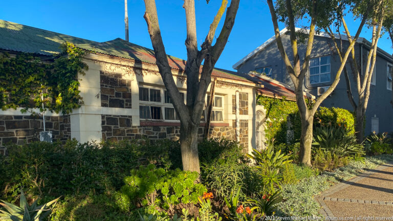 Low stone-and-plaster building with red brick trim, pitched roof, trees, and dense garden planting.