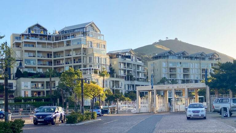 Pale apartment buildings with balconies beside a waterfront street, backed by the slope of Signal Hill.