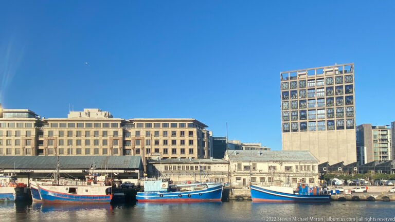 Several colorful working boats moored beside old harbor sheds, with the Zeitz MOCAA silo tower behind.