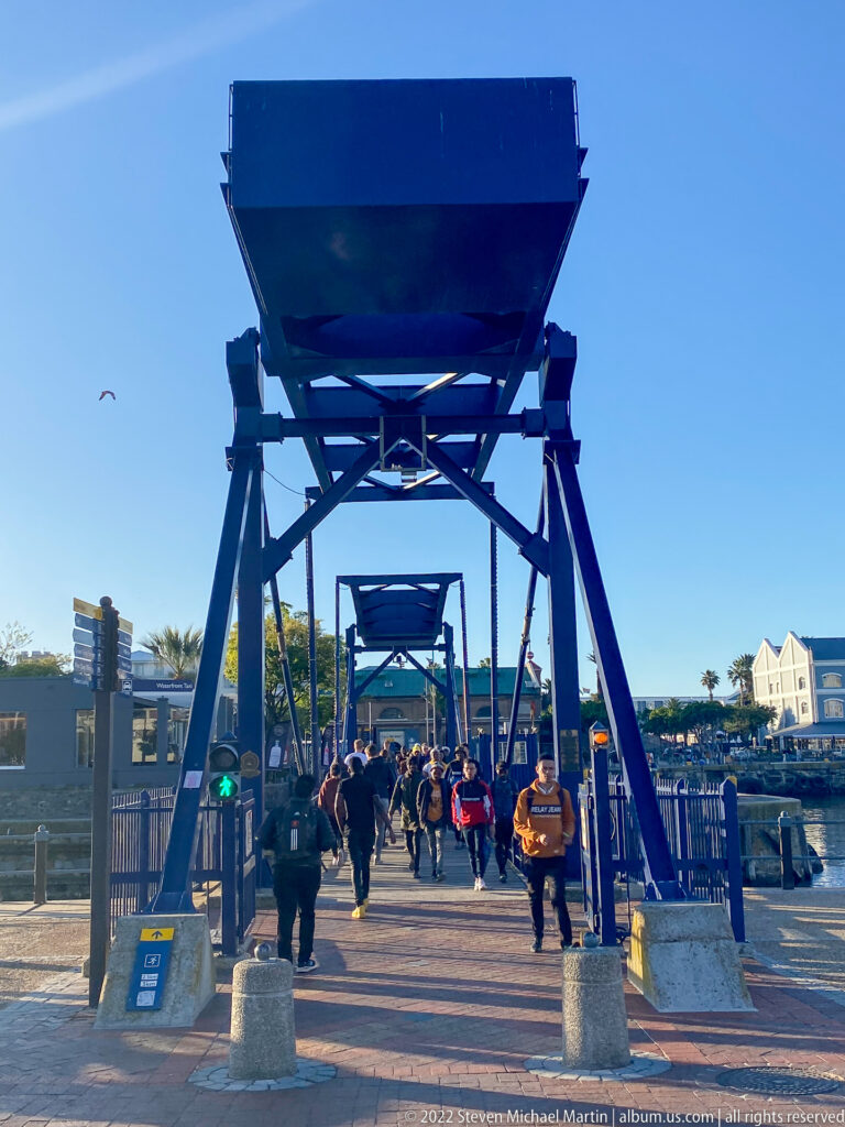 Pedestrians walking beneath a large blue steel movable bridge frame beside the harbor.