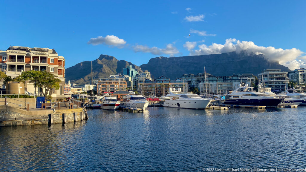 Yachts in a V&A Waterfront marina with apartment buildings and cloud-topped Table Mountain behind.