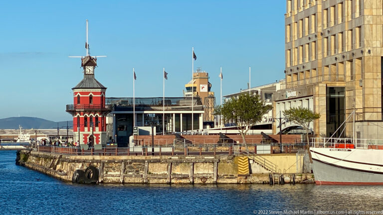 Red V&A Waterfront Clock Tower beside a harbor quay, modern buildings, flags, and moored boats.