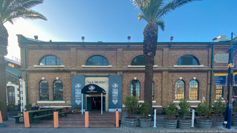 Brick V&A Food Market building with tall arched windows, entrance banners, palm tree, and outdoor seating.