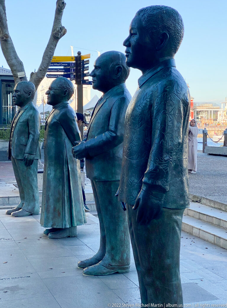 Four life-size bronze human figures standing in a row on a waterfront plaza.