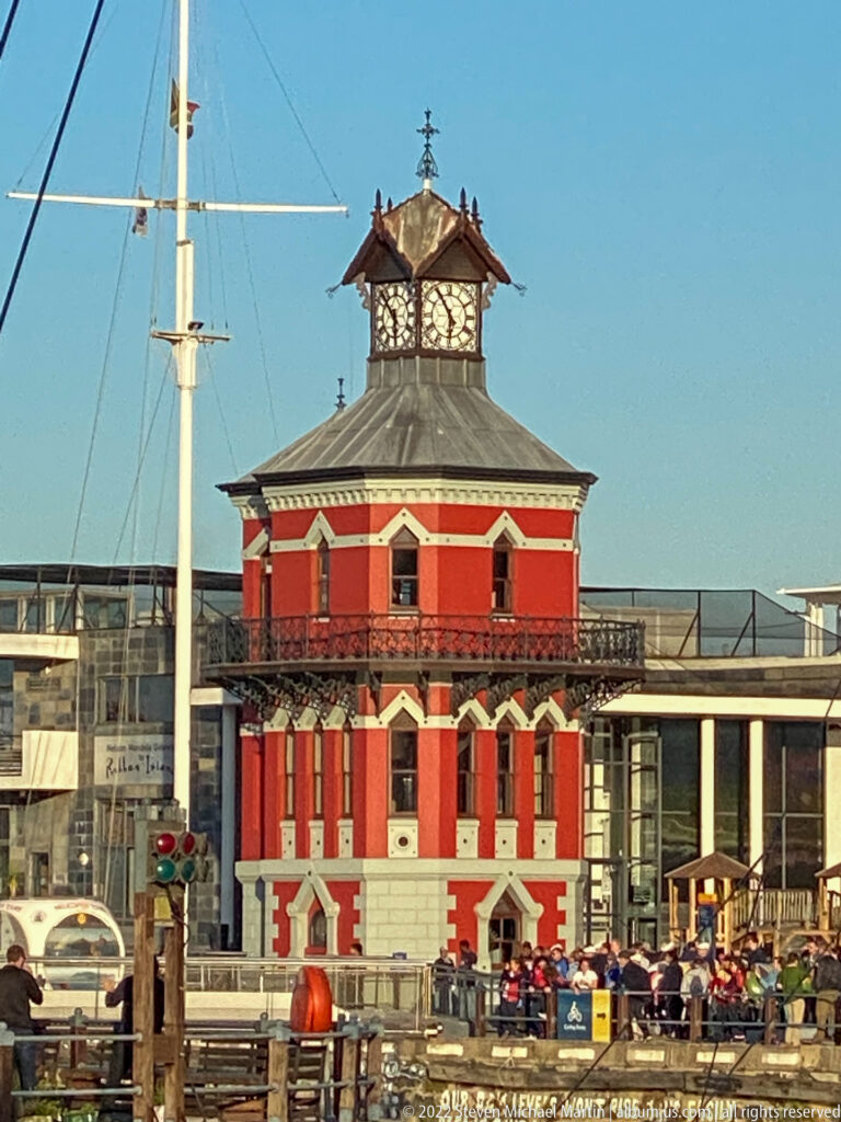Frontal view of the red V&A Waterfront Clock Tower with cream trim, balcony, roof, and upper pavilion.