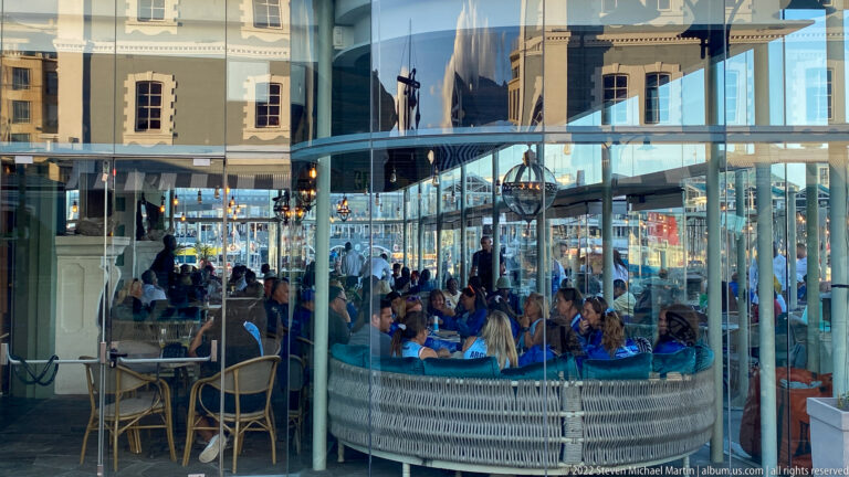 Crowded restaurant dining area behind glass, overlaid by reflections of nearby waterfront buildings.