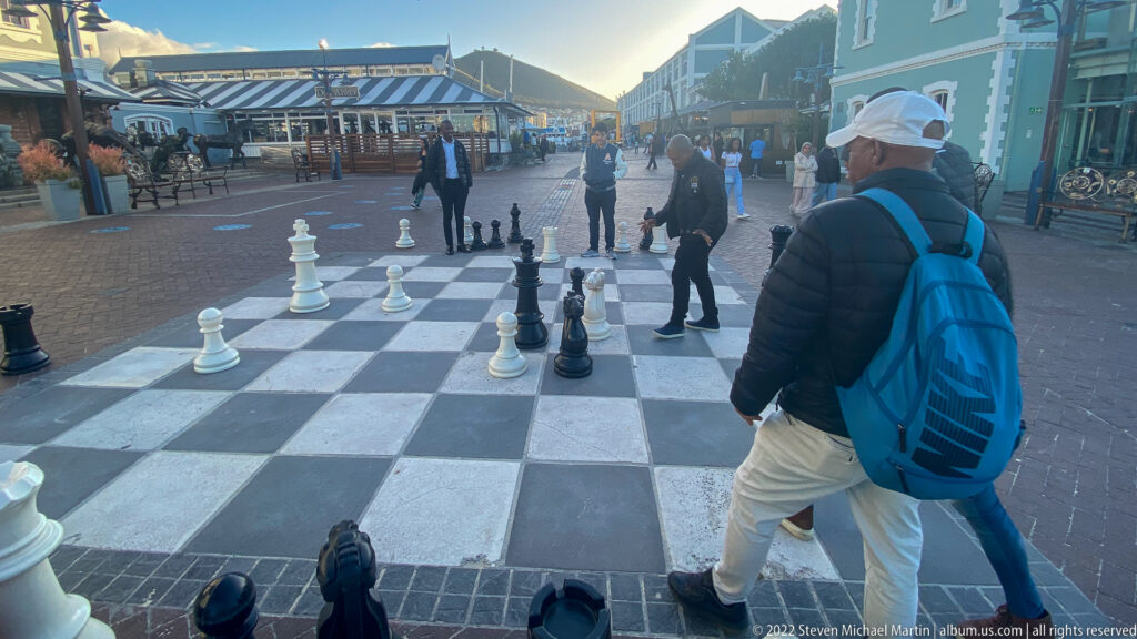Large outdoor chessboard with oversized black and white pieces and visitors walking around it.