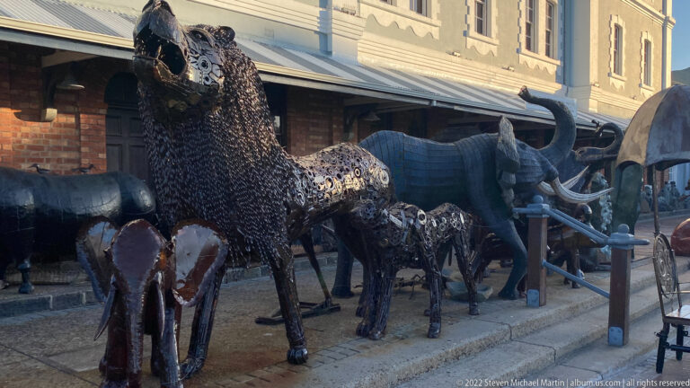 Group of welded dark-metal animal sculptures including giraffes and rhinoceroses outside waterfront shops.