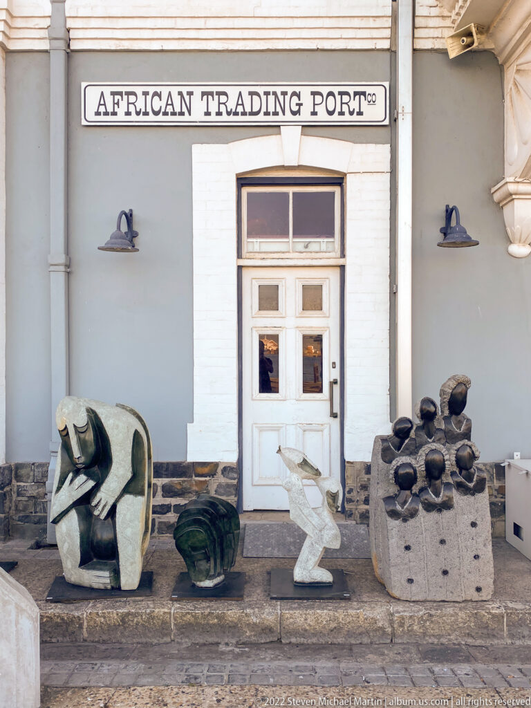Several sculptural objects displayed outside a pale building beneath the words African Trading Port.