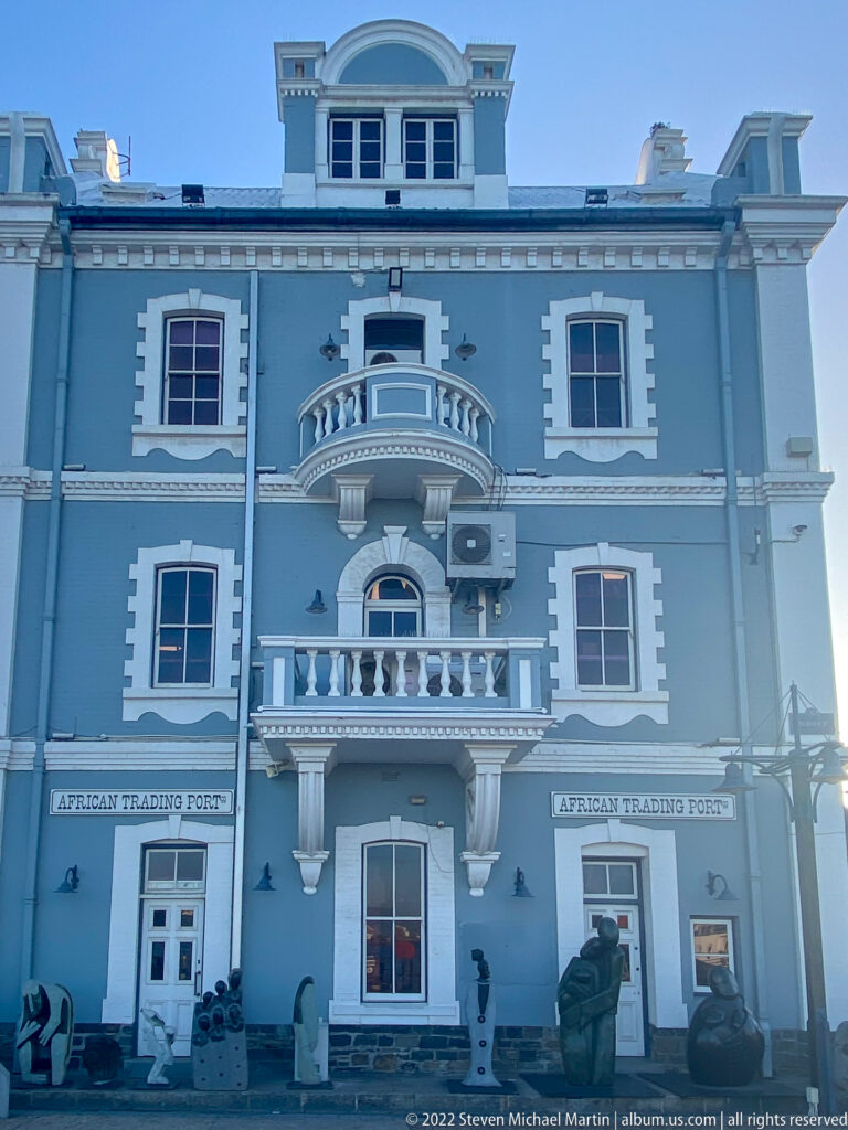 Pale blue African Trading Port façade with white trim, balconies, arched openings, and sculptures at street level.