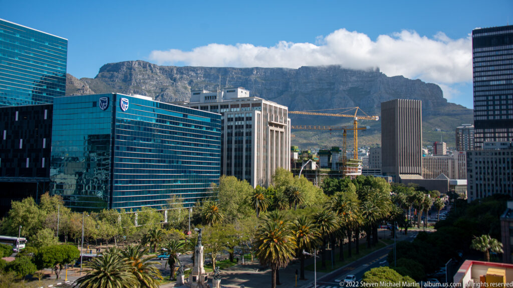 Table Mountain under low white cloud behind modern Cape Town office buildings, cranes, palms, and a city street.