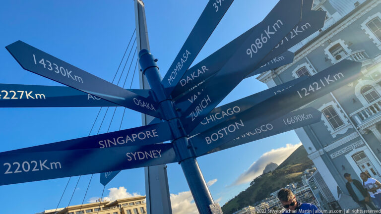 Dark radial signpost listing international cities and kilometer distances against blue sky and a mountain slope.