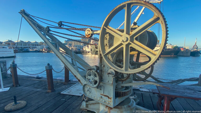 Historic quay crane with large spoked gear, chains, smaller gears, and angled boom beside harbor water.