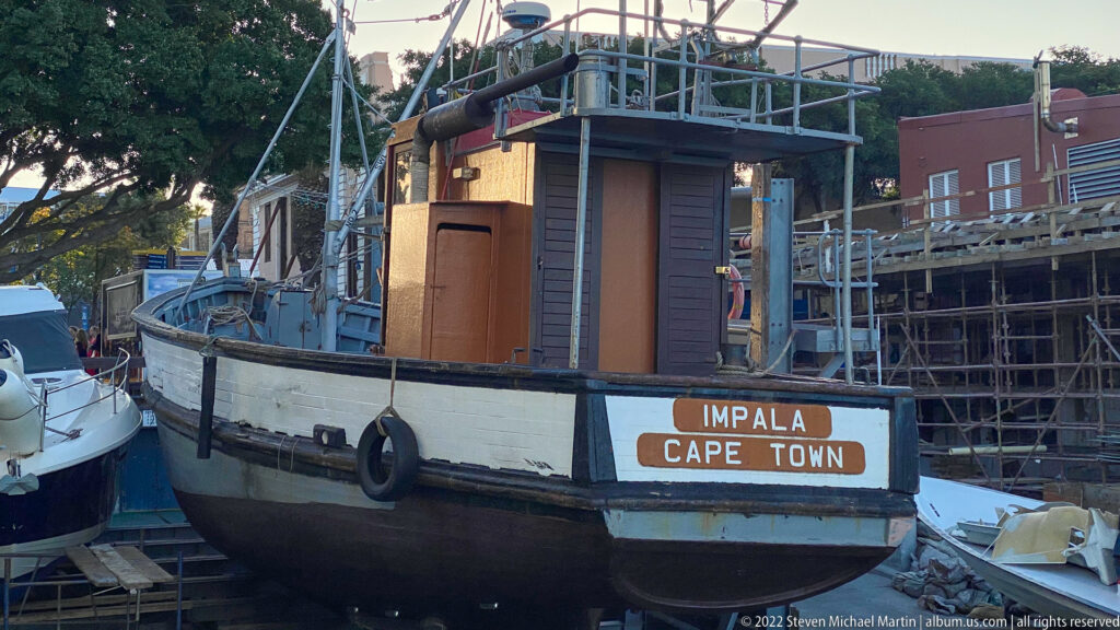 Stern of the vessel IMPALA marked CAPE TOWN, with timber sides, deckhouse, railings, and nearby boats.