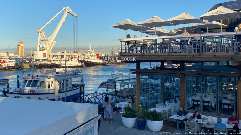 Harbor House restaurant terraces with white umbrellas above a harbor basin filled with boats and cranes.