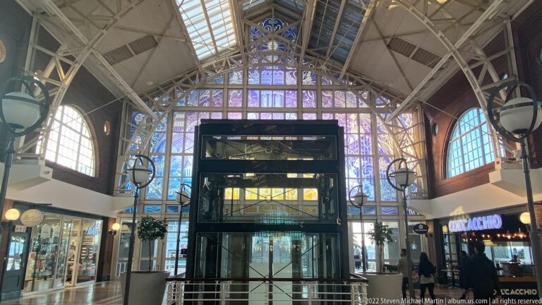 Glass elevator beneath a high iron-and-glass vaulted roof with arched windows inside Victoria Wharf.