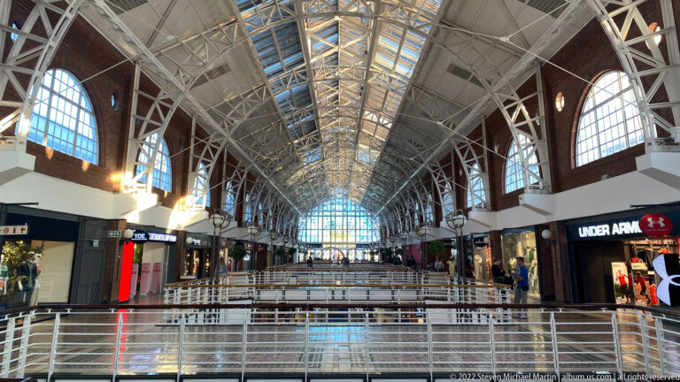 Long shopping arcade with glazed barrel roof, white trusses, arched windows, brick walls, and storefronts.