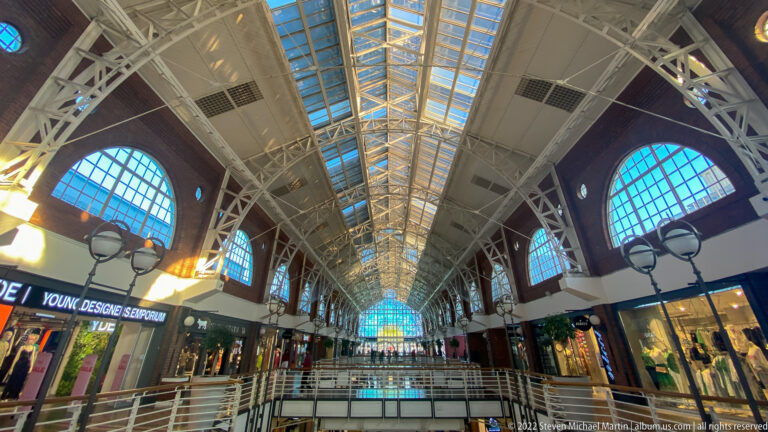 Upward view of white roof trusses, glazed skylights, and large arched windows above Victoria Wharf shops.