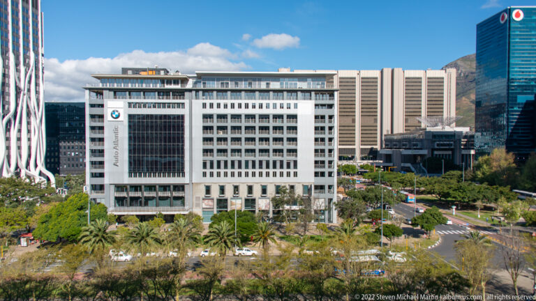 White Allan Gray office building beyond palms and a landscaped boulevard, with modern towers and a mountain slope at right.
