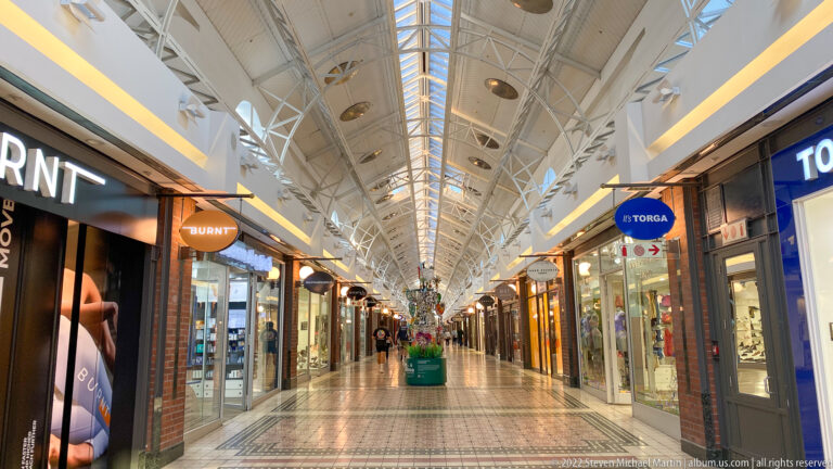 Long retail corridor with glass storefronts, patterned floor, white trusses, skylights, and distant shoppers.