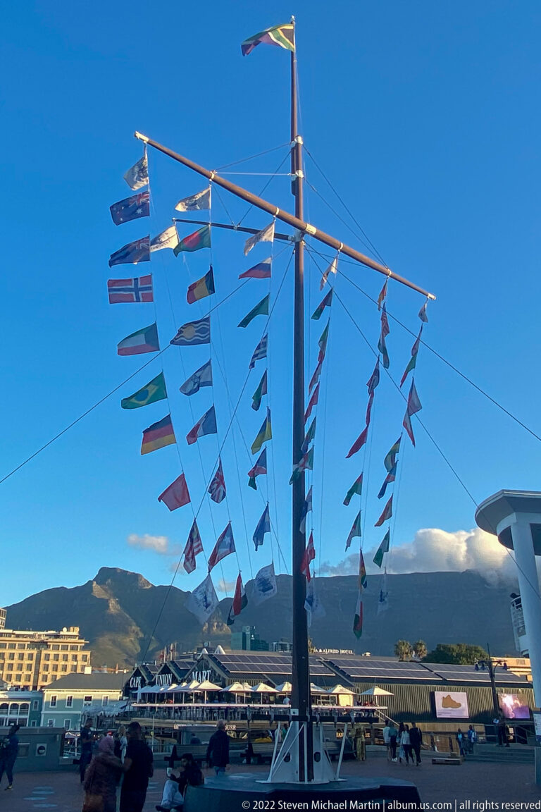 Tall mast hung with many colorful flags above the waterfront, with Table Mountain in the background.
