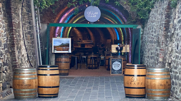 Stone-vaulted entrance to The Cellar Club with rainbow lighting, wine barrels, stools, and signage.