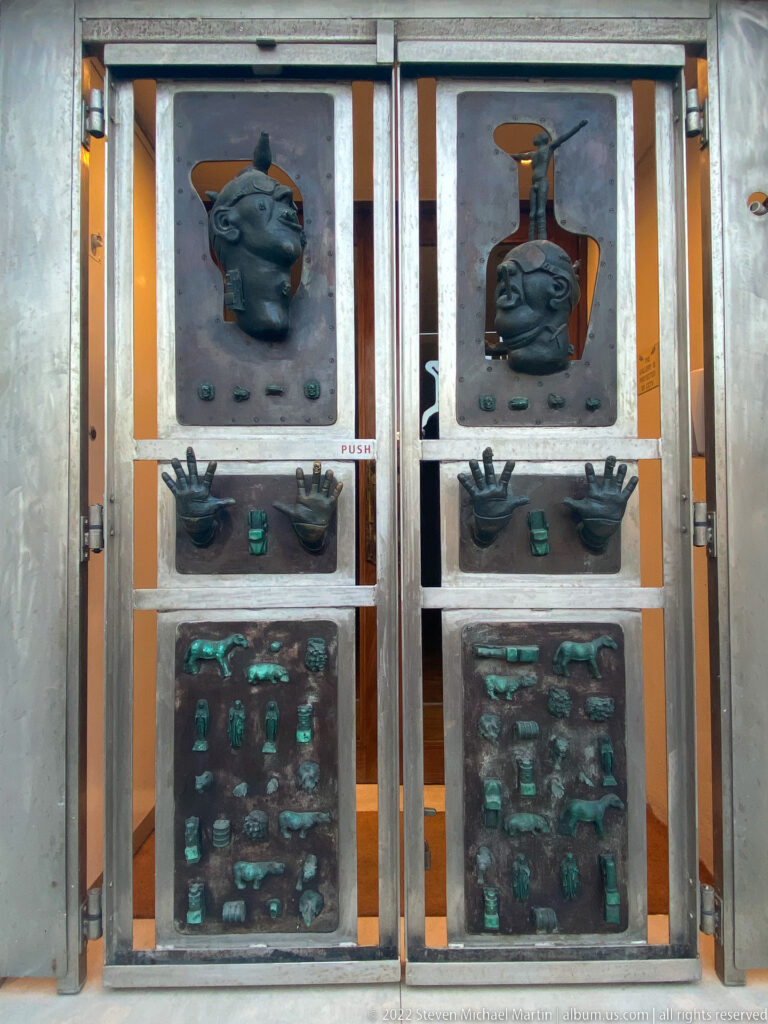Paired metal doors with raised human heads, hands, and rows of small green-patinated decorative motifs.
