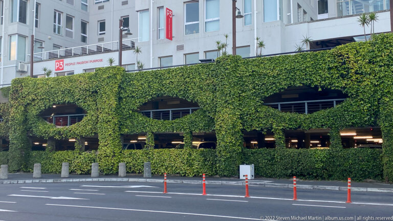 Multi-story parking structure covered in dense green ivy, with oval openings and roadway in front.