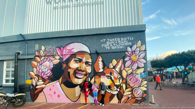 Colorful mural of a smiling person, antelope, and flowers beside the words about sun and rain.