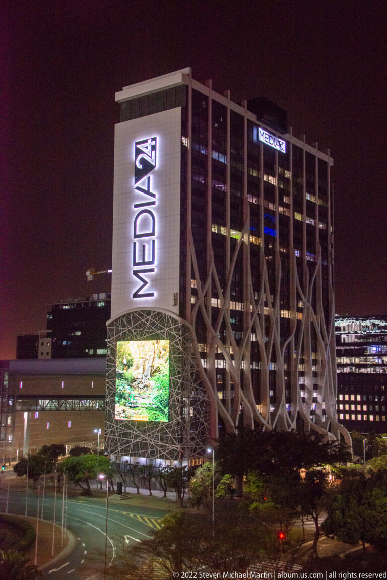 Media24 office building at night with illuminated vertical MEDIA24 signage, branching supports, and a bright digital screen.