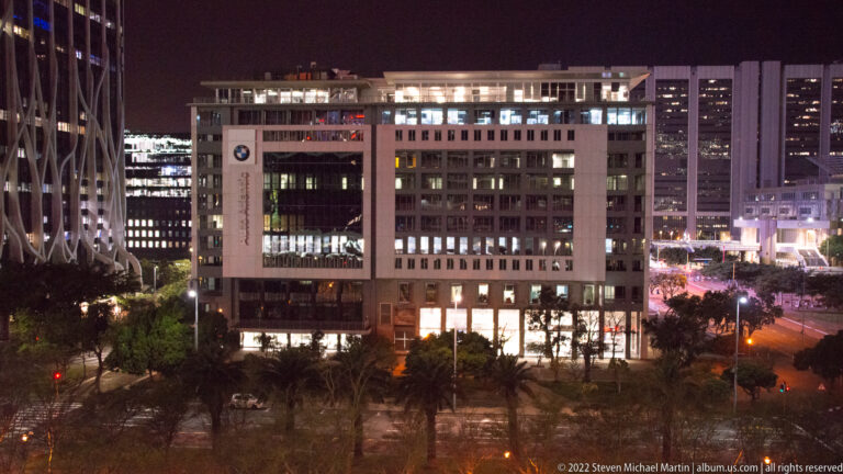 Modern office building at night with illuminated windows, a large central opening, trees, and neighboring towers.