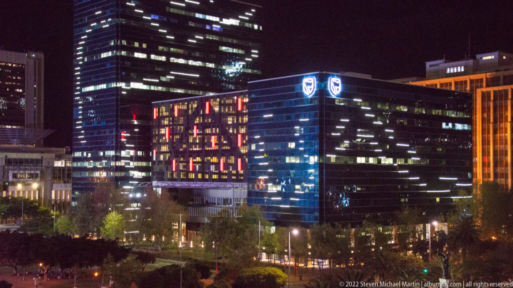 Dark glass office towers at night with lit windows, corporate logos, and a colorful illuminated façade panel.