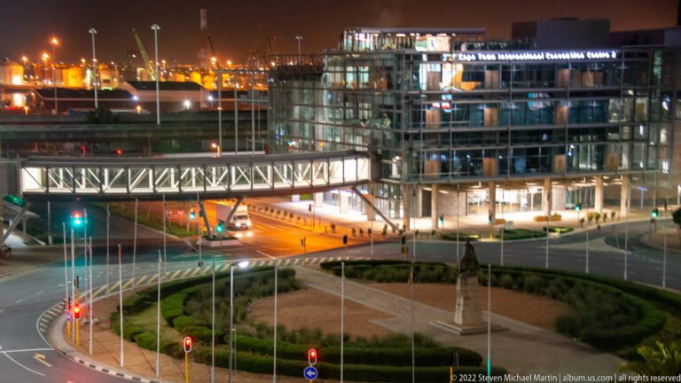 Night view of a landscaped roundabout, flagpoles, pedestrian bridge, traffic lights, and modern office buildings.