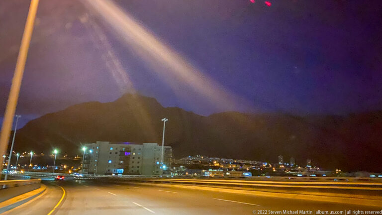 Curving Cape Town roadway at night with city lights and the dark silhouette of Table Mountain ahead.