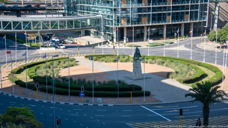 Landscaped roundabout with a standing monument at center, surrounded by multilane roads, glass buildings, and a pedestrian bridge.