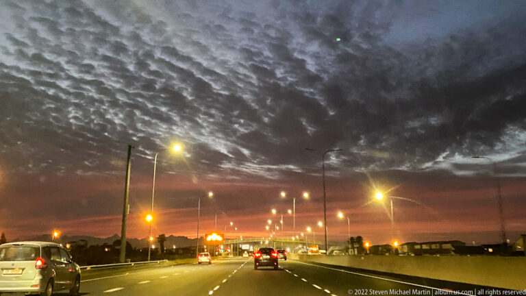 Cars traveling on a multi-lane roadway at night beneath rippled clouds and rows of bright streetlights.