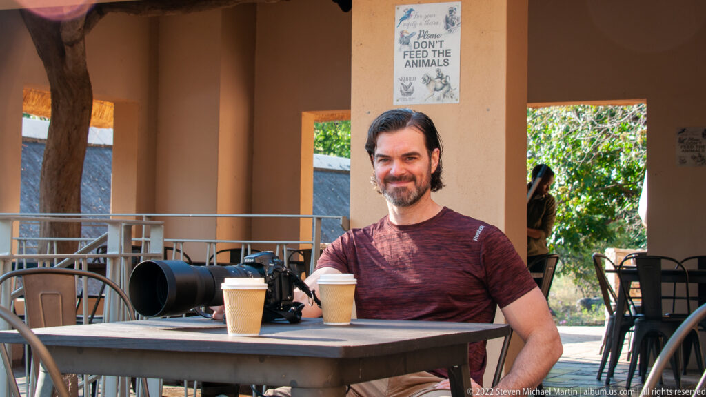Man seated at a shaded outdoor café table with two drinks and camera gear beside him.