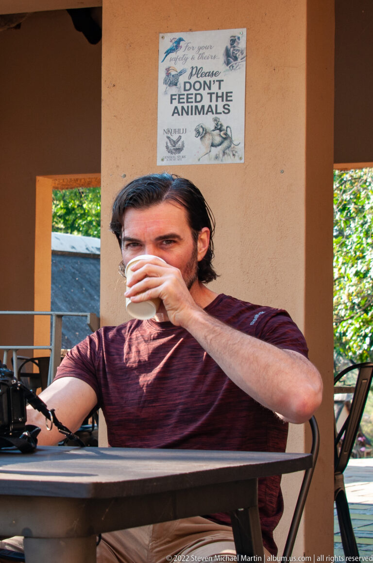 Seated man drinking from a takeaway cup at an outdoor table with camera equipment in front of him.