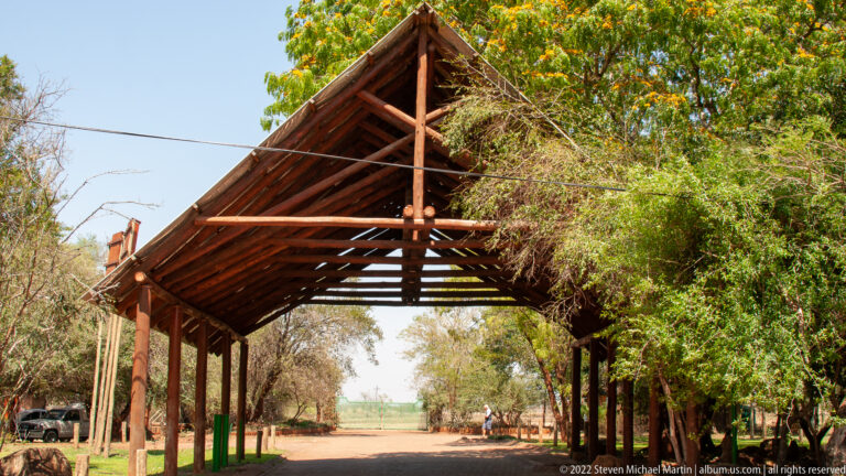 Tall timber gateway with steep pitched roof and exposed trusses spanning a road through trees.