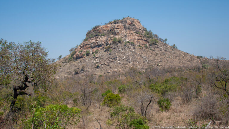 Rocky rounded hill with exposed boulders rising above dry woodland under a clear blue sky.